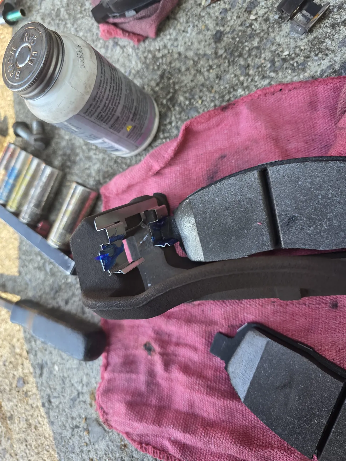 New brake pads with their clips on a red shop rag, beside a can of synthetic caliper grease and a row of sockets
