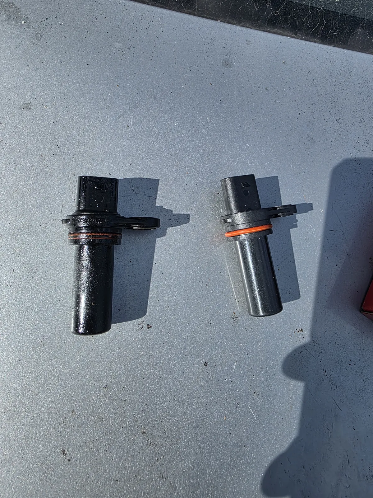 An old, oil-blackened engine position sensor next to its new replacement with a fresh orange O-ring