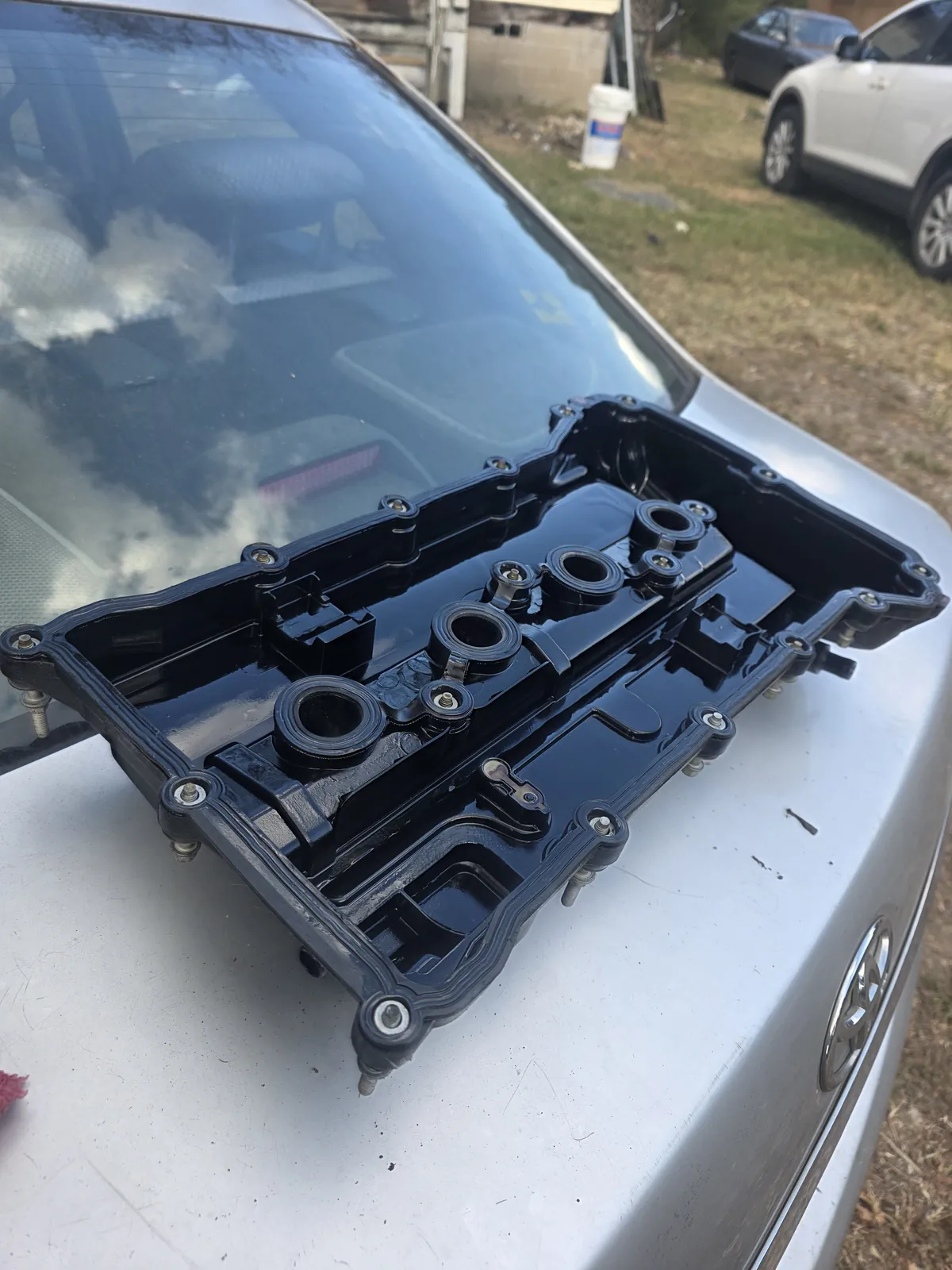 A black valve cover off the engine, resting on the hood of a silver Toyota parked in a yard